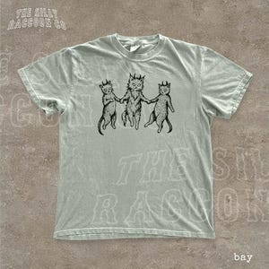 A light green t-shirt with a graphic design featuring three cats with horns and tails, holding hands and appearing to dance or interact with each other.