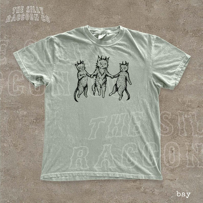 A light green t-shirt with a graphic design featuring three cats with horns and tails, holding hands and appearing to dance or interact with each other.