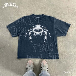 A navy blue t-shirt with a white spider graphic printed on it, laid out on a concrete floor. A pair of white sneakers is visible underneath the t-shirt.