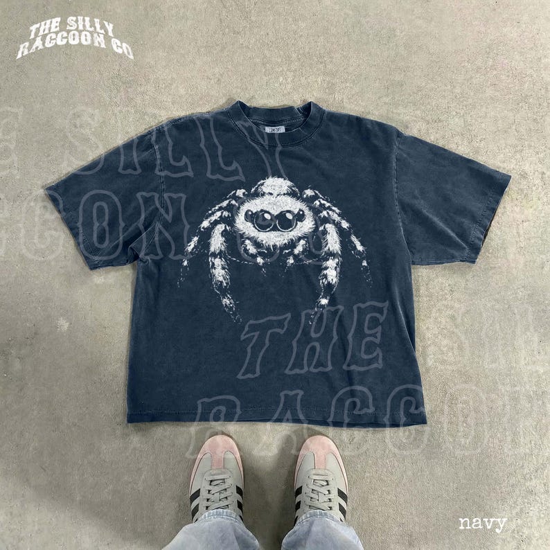 A navy blue t-shirt with a white spider graphic printed on it, laid out on a concrete floor. A pair of white sneakers is visible underneath the t-shirt.