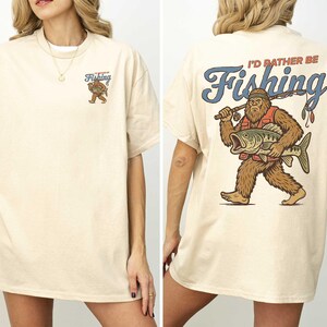 a woman wearing a white t-shirt with a graphic design on the back. The design depicts a large fish with a bear-like appearance, and the text on the back reads "I'd rather be Fishing".