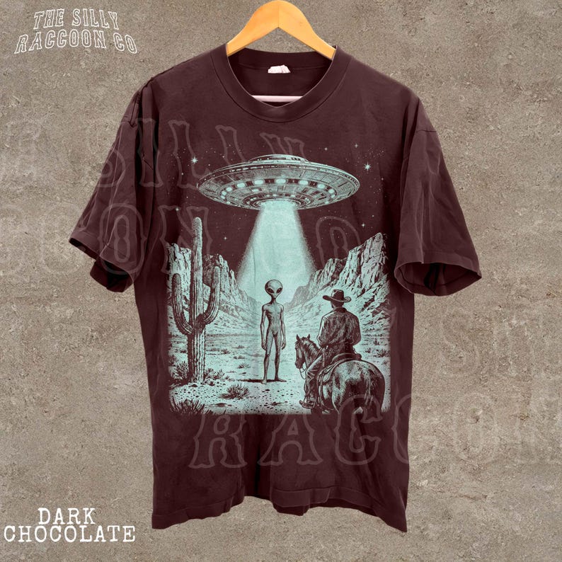 a t-shirt with a graphic design depicting an alien spacecraft hovering over a desert landscape with a cowboy on a horse.