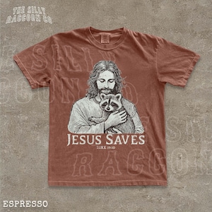 A t-shirt with a graphic design depicting a man holding a raccoon, along with the text "JESUS SAVES" and "LIKE 19:10".