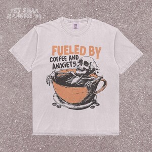 Funny coffee shirt for anxious introverts, featuring a skeleton relaxing in a giant coffee cup with the text “Fueled by Coffee and Anxiety.” Vintage grunge design with distressed print and muted tones. Ideal for women, men, and non-binary adults who love dark humor and caffeine culture. Great for Halloween, work-from-home vibes, mental health awareness, or gifting your favorite goth coffee lover.