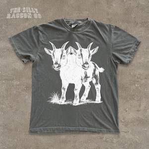 May include: Dark gray t-shirt with a white illustration of a two-headed goat. The text "THE SILLY RACCOON CO" is printed at the top. The shirt is made of a soft material and has a crew neck.
