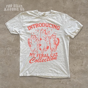 A white t-shirt with a graphic design featuring three cats and the text "INTRODUCING MY FERAL CAT COLLECTION" printed on it.