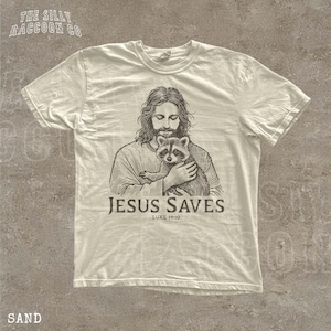 A t-shirt with a graphic design depicting a man holding a raccoon, along with the text "JESUS SAVES" and "LUKELI 19:10".