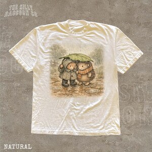 A white t-shirt with a printed illustration of two hedgehog characters sharing an umbrella in the rain.