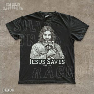 A black t-shirt with a graphic design depicting a man holding a raccoon. The text below the graphic reads "JESUS SAVES LUKE 19:10".