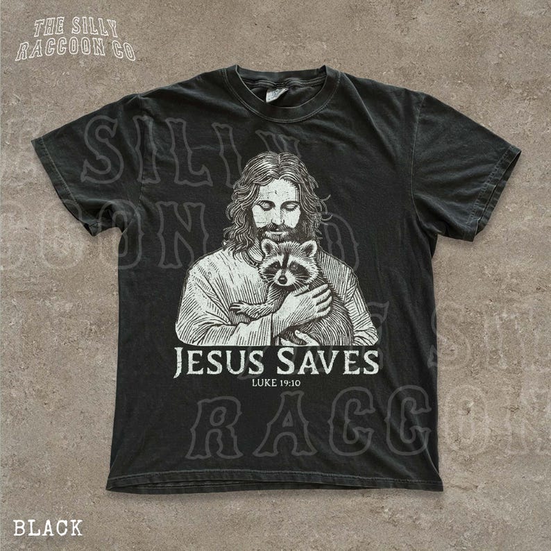 A black t-shirt with a graphic design depicting a man holding a raccoon. The text below the graphic reads "JESUS SAVES LUKE 19:10".