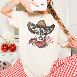 A person is taking a selfie with a camera, wearing a white t-shirt with a graphic of a dog wearing a cowboy hat and a red scarf.