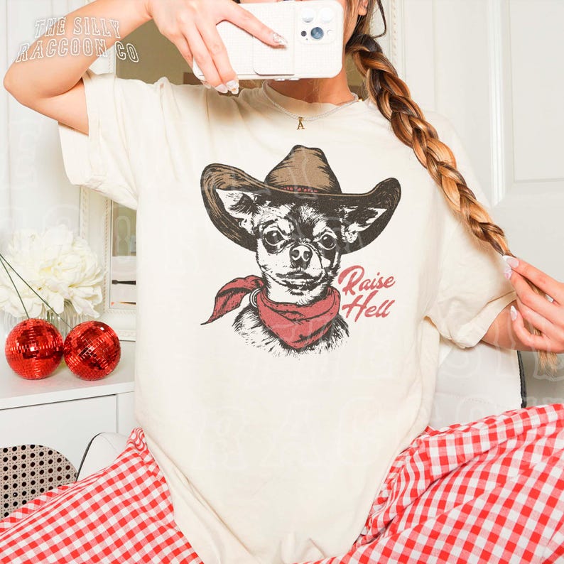 A person is taking a selfie with a camera, wearing a white t-shirt with a graphic of a dog wearing a cowboy hat and a red scarf.
