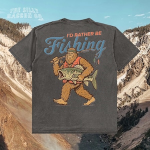 a t-shirt with a graphic design depicting a large, furry creature holding a fish and walking on all fours, with mountains in the background.