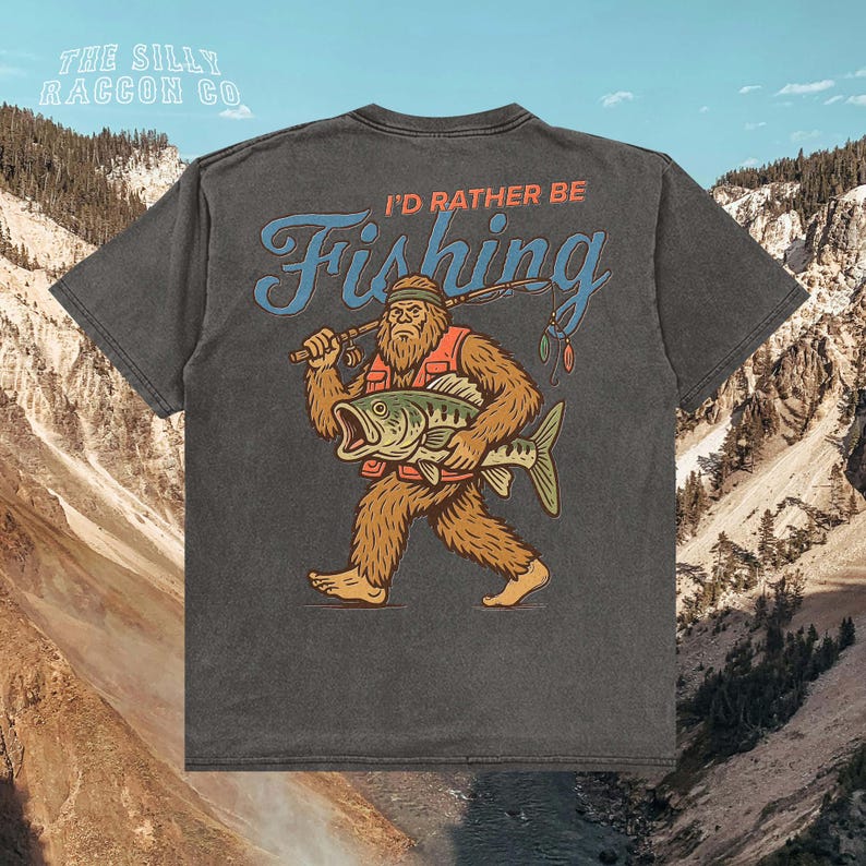 a t-shirt with a graphic design depicting a large, furry creature holding a fish and walking on all fours, with mountains in the background.