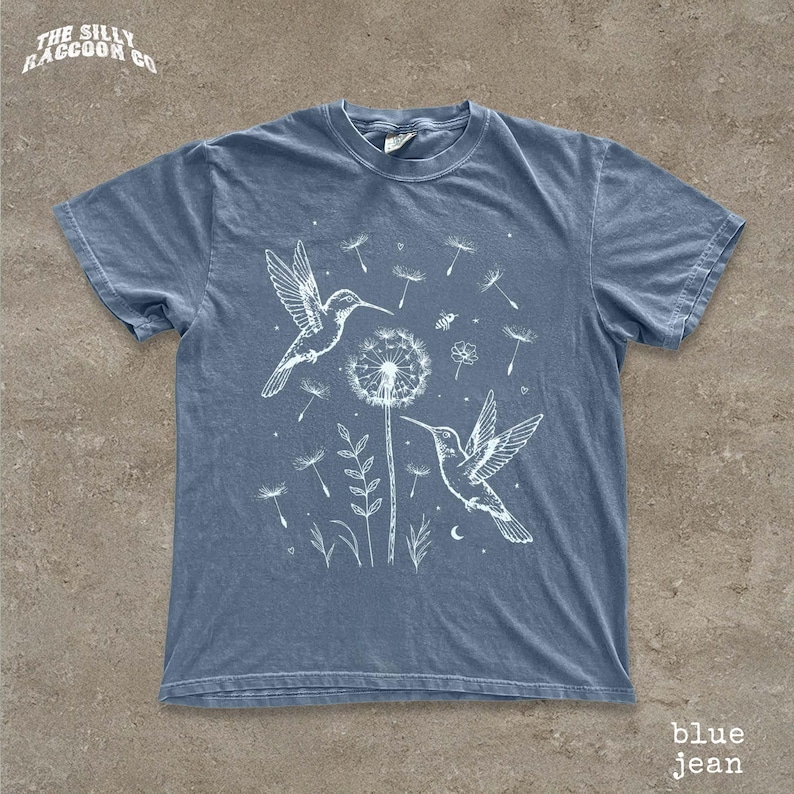 A blue t-shirt with a white illustration of two hummingbirds and a dandelion on a flower, along with other floral and bird-like elements.