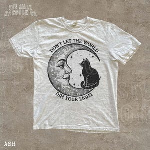 a white t-shirt with a graphic design depicting a cat sitting on a crescent moon and the text "Don't Let the World Dim Your Light" printed on it.