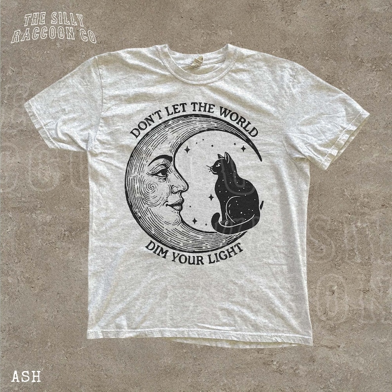 a white t-shirt with a graphic design depicting a cat sitting on a crescent moon and the text "Don't Let the World Dim Your Light" printed on it.