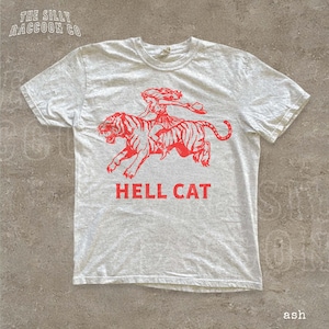 A white t-shirt with a graphic design depicting a person riding a tiger, accompanied by the text "HELL CAT" in red letters.
