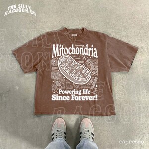 A brown t-shirt with a graphic design depicting Mitochondria, a cellular organelle, and the text "Powering life since forever!" printed on it. The t-shirt is laid out on a concrete floor, and a pair of feet is visible in the bottom right corner of the image.