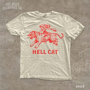 A white t-shirt with a graphic design depicting a person riding a tiger, accompanied by the text "HELL CAT" in red letters.