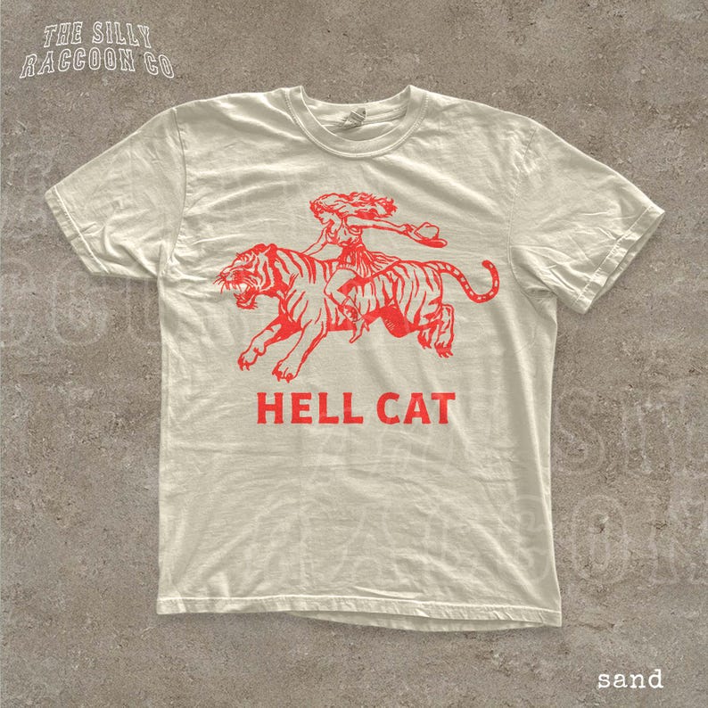 A white t-shirt with a graphic design depicting a person riding a tiger, accompanied by the text "HELL CAT" in red letters.