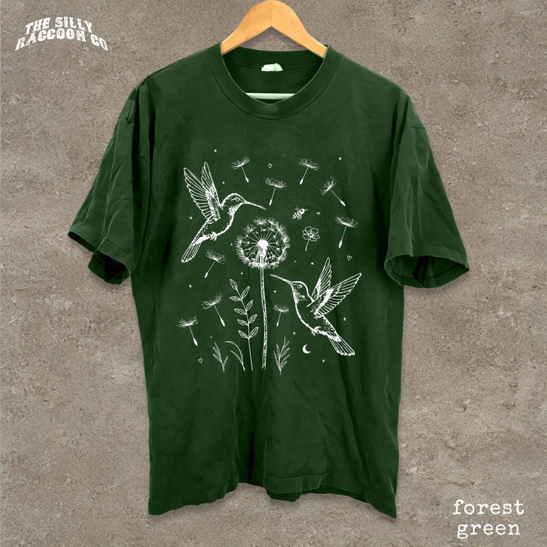 A green t-shirt with a white illustration of two hummingbirds hovering over a dandelion surrounded by flowers and plants.
