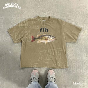 A pair of sneakers and a t-shirt with a fish graphic are shown on a concrete floor.