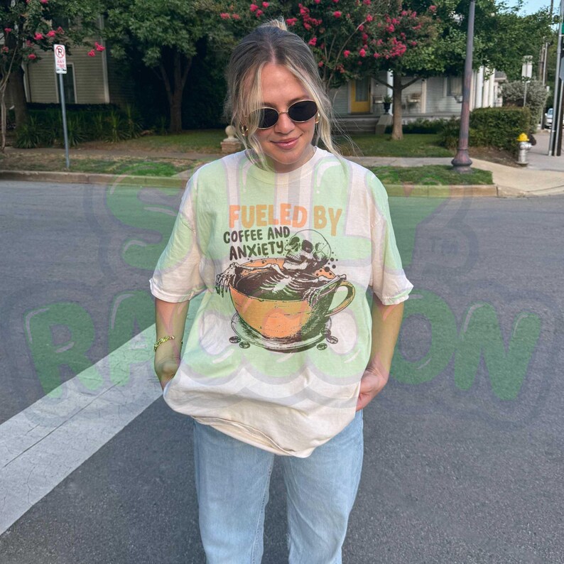 Funny coffee shirt for anxious introverts, featuring a skeleton relaxing in a giant coffee cup with the text “Fueled by Coffee and Anxiety.” Vintage grunge design with distressed print and muted tones. Ideal for women, men, and non-binary adults who love dark humor and caffeine culture. Great for Halloween, work-from-home vibes, mental health awareness, or gifting your favorite goth coffee lover.