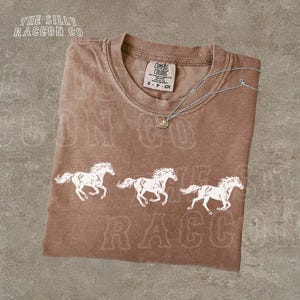 A brown t-shirt with a graphic design of three white horses running, along with text that reads "The Silly Raccoon Co."