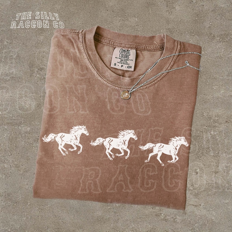 A brown t-shirt with a graphic design of three white horses running, along with text that reads "The Silly Raccoon Co."