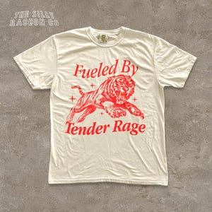 A white t-shirt with a red graphic design depicting a tiger and the text "Fuelled By Tender Rage" printed on it.