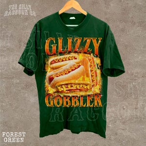 A green t-shirt with a graphic design featuring two hot dogs on a bun, with flames or fire around them, and the text "GLIZZY GOBBLER" prominently displayed.