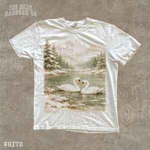 A white t-shirt with a printed image of two swans swimming in a lake, surrounded by snow-covered trees and mountains.