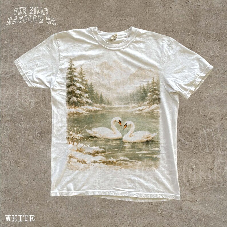 A white t-shirt with a printed image of two swans swimming in a lake, surrounded by snow-covered trees and mountains.