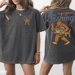 a woman wearing a grey t-shirt with a graphic design on the back. The design depicts a man holding a fishing rod and a fish, with the text "I'd rather be fishing" written above the image.
