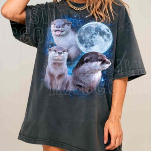 A woman wearing a black t-shirt with a graphic design featuring three otters against a backdrop of a full moon and stars.