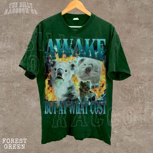 A green t-shirt with a graphic design featuring two koalas and the text "AWAKE BUT AT WHAT COST" printed on it.