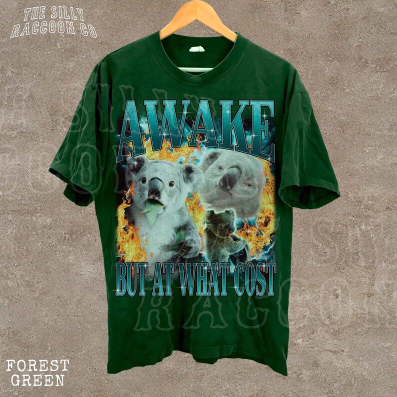 A green t-shirt with a graphic design featuring two koalas and the text "AWAKE BUT AT WHAT COST" printed on it.