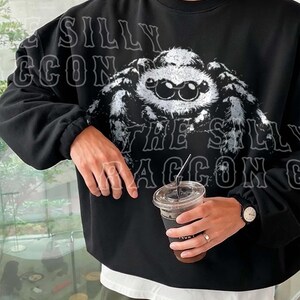 A person wearing a black sweatshirt with a large spider graphic on it, holding a drink in their hand.