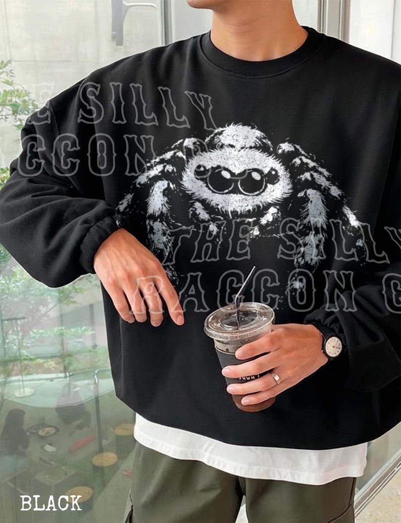 A person wearing a black sweatshirt with a large spider graphic on it, holding a drink in their hand.