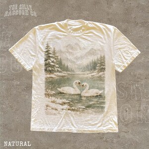 A white t-shirt with a printed image of two swans swimming in a river, surrounded by snow-covered trees.