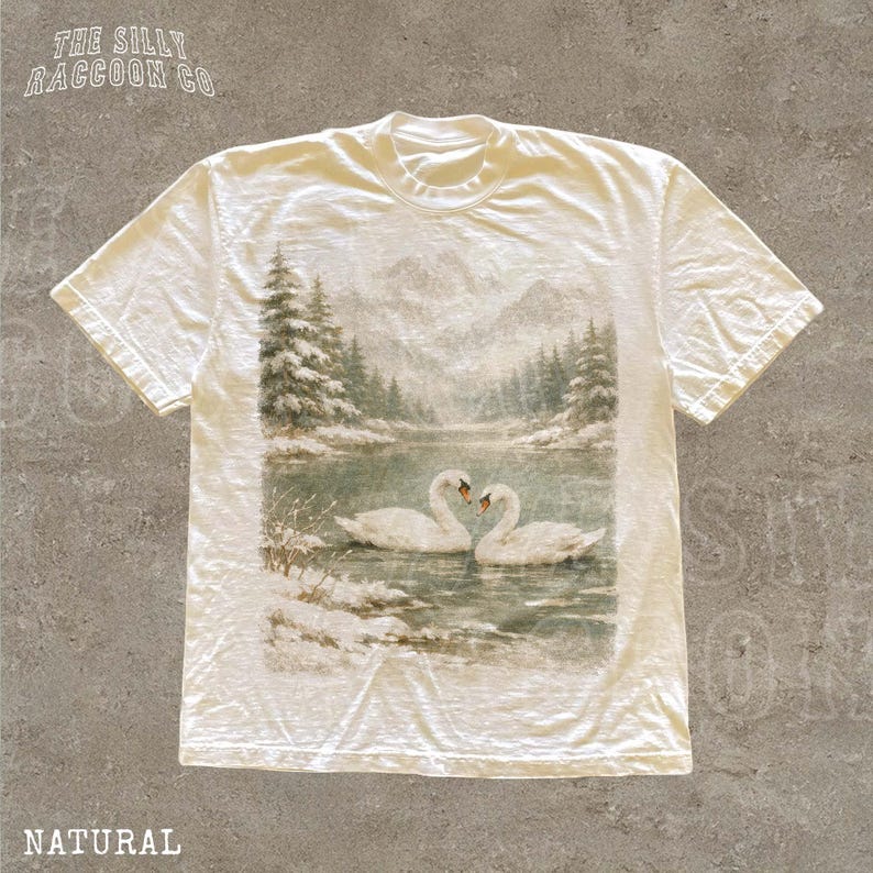 A white t-shirt with a printed image of two swans swimming in a river, surrounded by snow-covered trees.