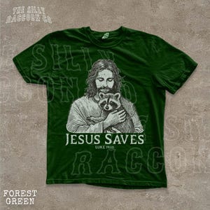 A green t-shirt with a graphic design depicting a man holding a raccoon. The text below the image says "JESUS SAVES" and "LIKE YOU".