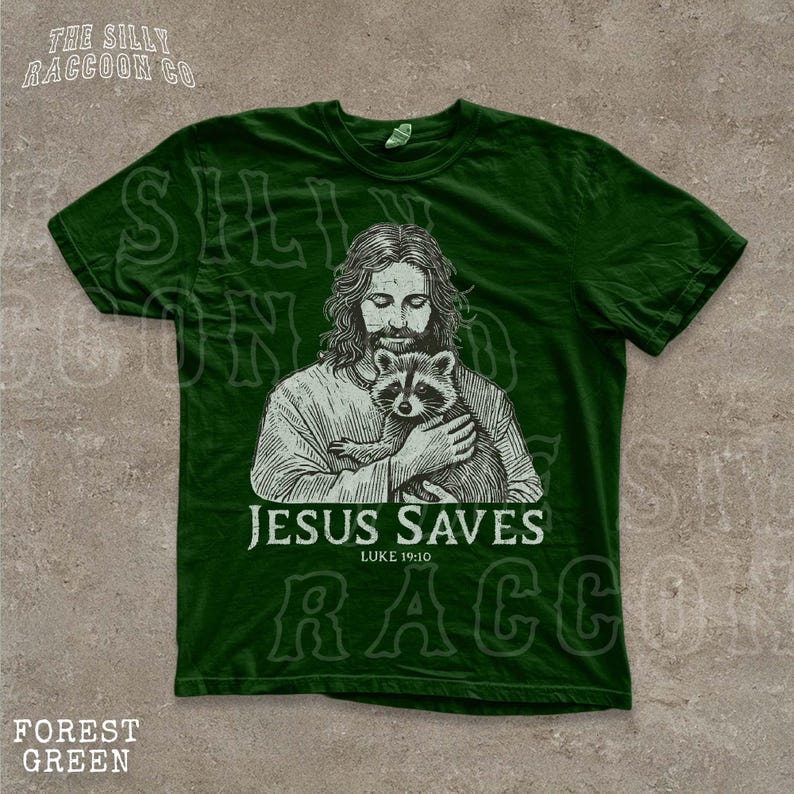 A green t-shirt with a graphic design depicting a man holding a raccoon. The text below the image says "JESUS SAVES" and "LIKE YOU".