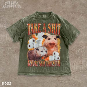 A t-shirt with a graphic design featuring an opossum surrounded by toilet paper rolls, with the text "TAKE A SHIT BEFORE SHIT TAKES YOU" prominently displayed.