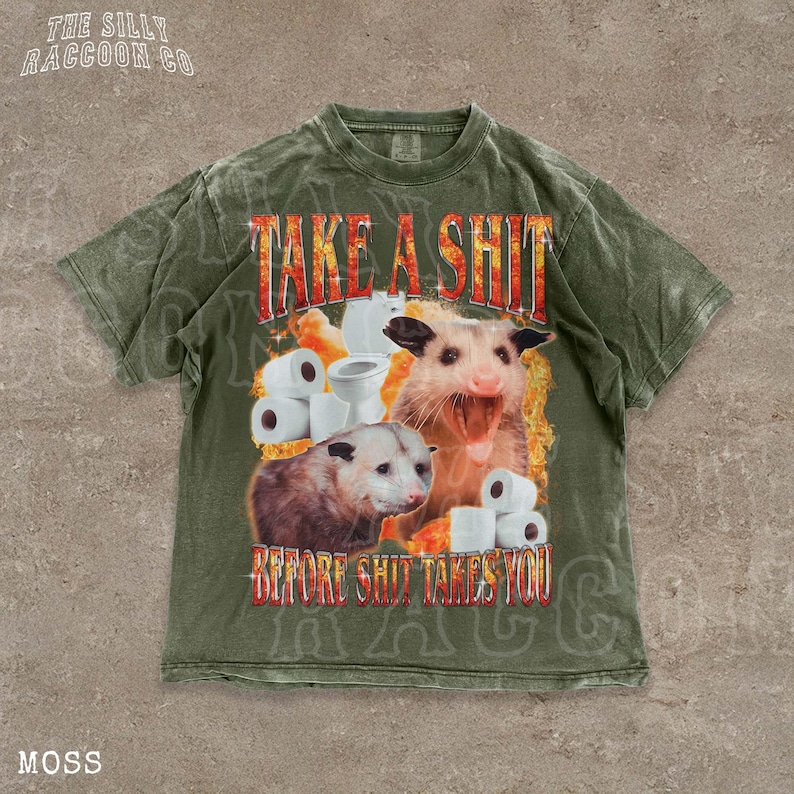 A t-shirt with a graphic design featuring an opossum surrounded by toilet paper rolls, with the text "TAKE A SHIT BEFORE SHIT TAKES YOU" prominently displayed.