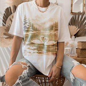 A person is sitting on a chair wearing a white t-shirt with a printed image of two swans on a lake.