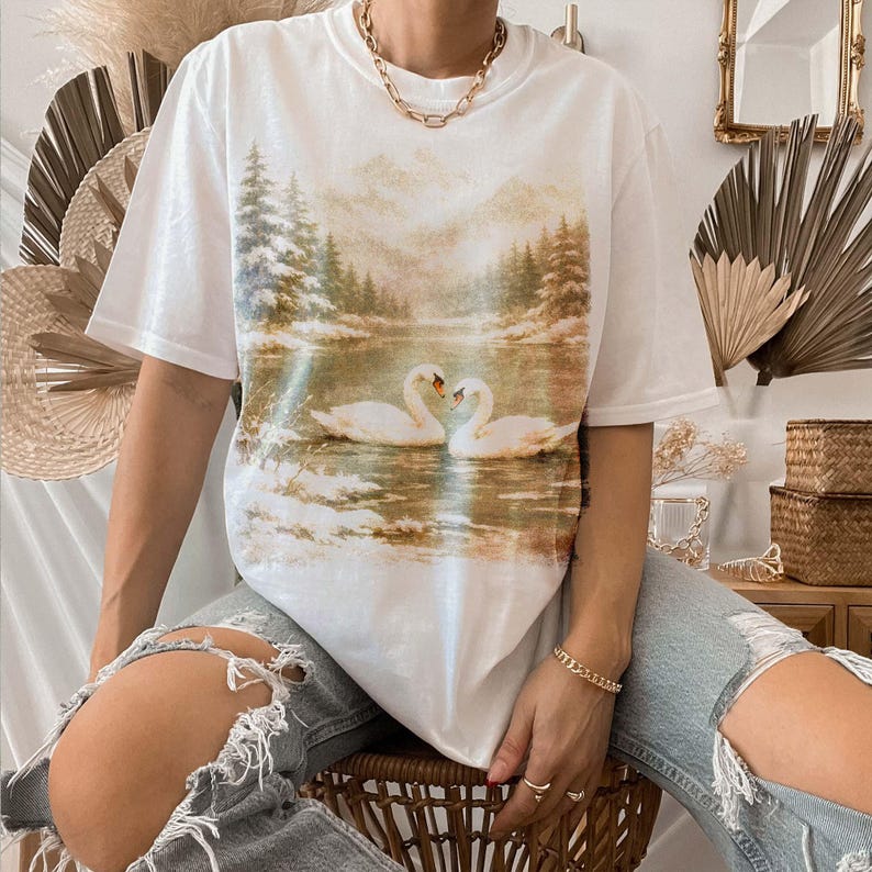 A person is sitting on a chair wearing a white t-shirt with a printed image of two swans on a lake.
