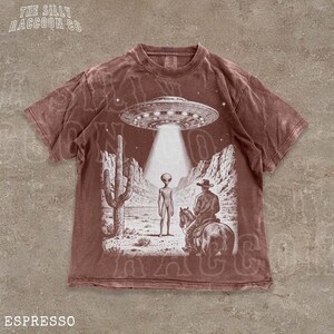 The image is a t-shirt design featuring an illustration of an alien spacecraft hovering over a desert landscape, with a cowboy on a horse below.