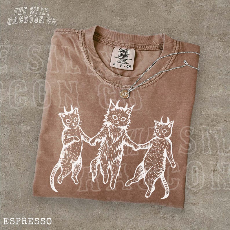 A brown t-shirt with a white illustration of three cats holding hands, accompanied by the text "ESPRESSO" and a tag that reads "COMFORT COLORS".
